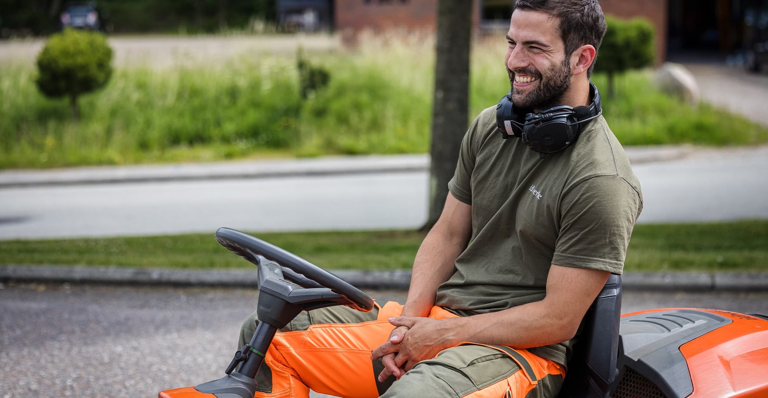 idverde worker smiling on orange ride-on mower, wearing headphones around neck, outdoors in green surroundings.
