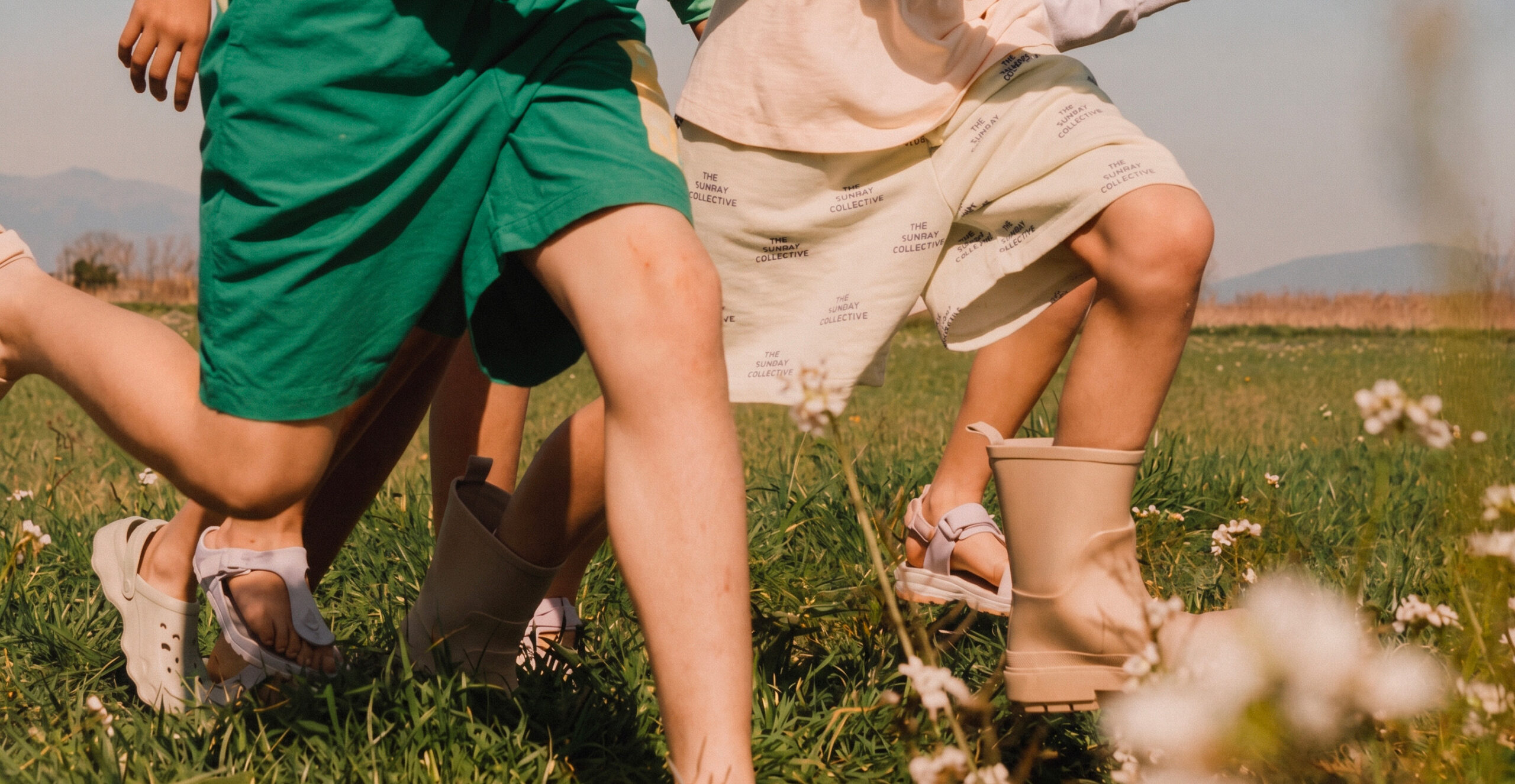 Children run barefoot through a wildflower meadow in summer, wearing casual clothing, with rolling hills visible in the background.