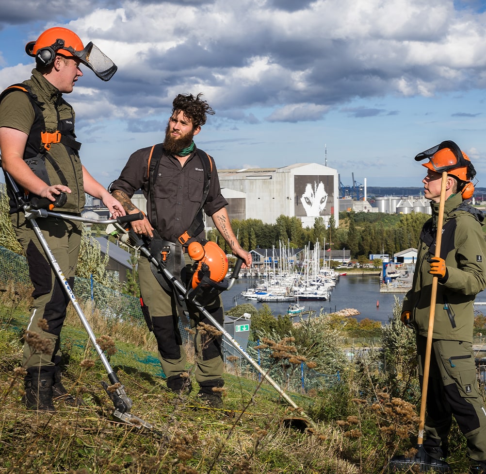 Three idverde grounds maintenance workers with safety helmets and tools on a hillside overlooking a marina and industrial port.