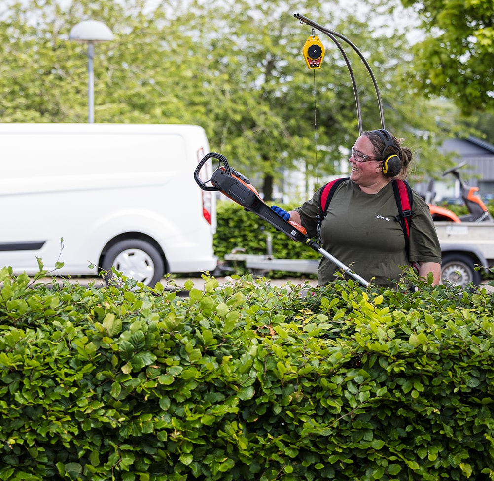 idverde worker trimming a green hedge with a powered hedge cutter and overhead tool balancer outdoors