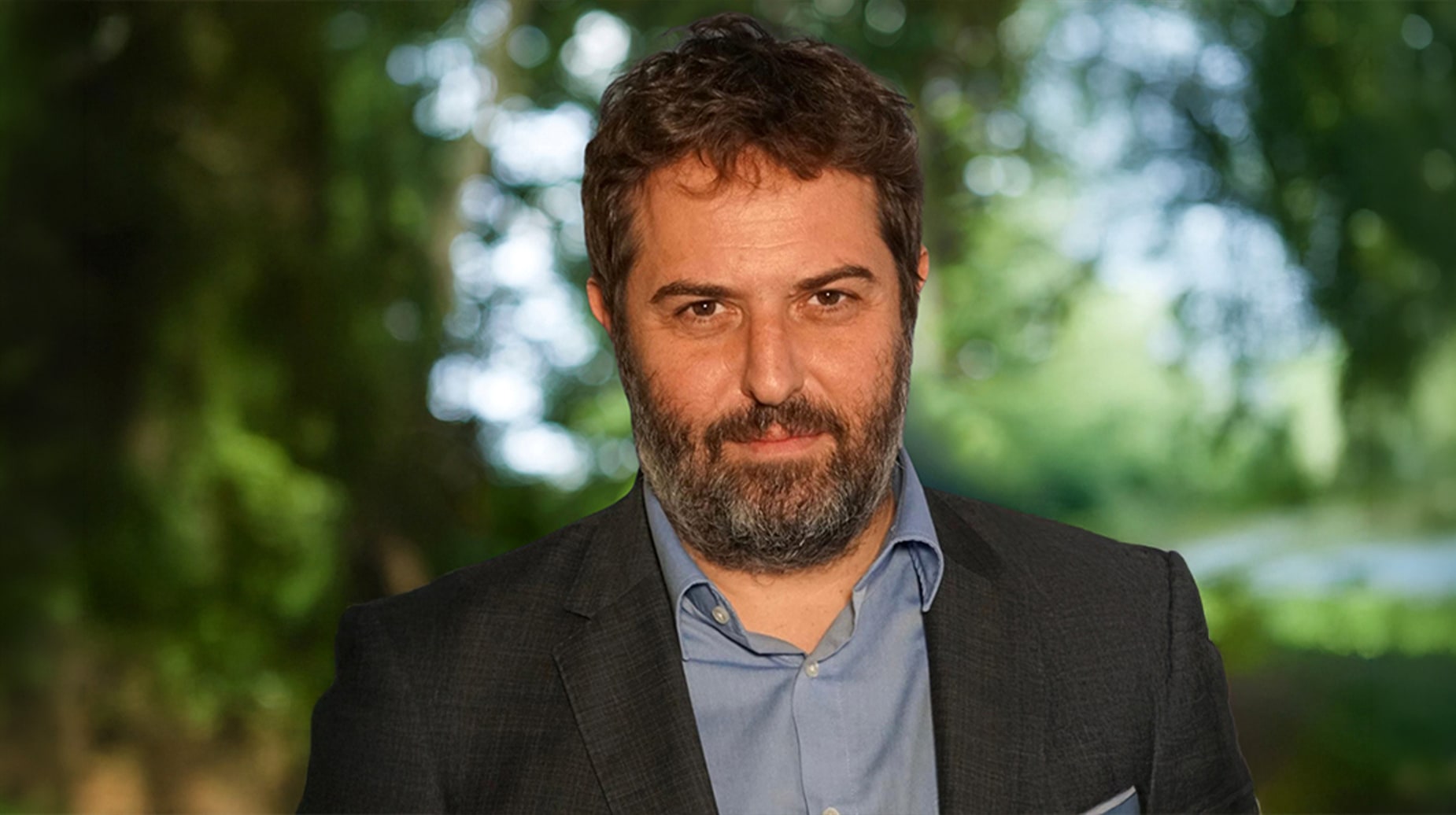 Photo of Lionel Beyllier wearing a grey blazer and blue shirt, photographed outdoors with blurred green trees.