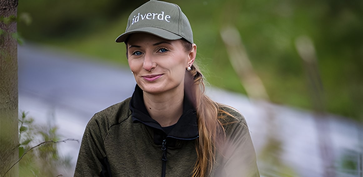 idverde employee smiling outdoors wearing a branded olive green cap and jacket in a natural green setting