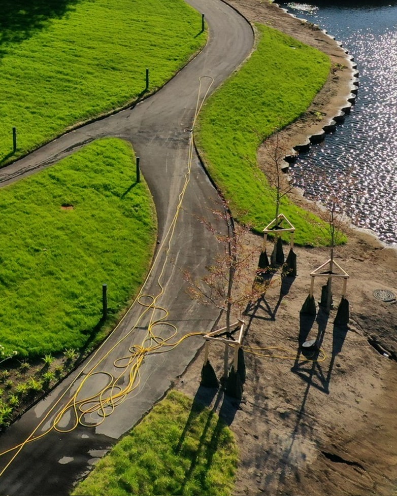 Aerial view of a curved path beside a reservoir, with newly planted trees supported by stakes and a yellow hose coiled on the ground
