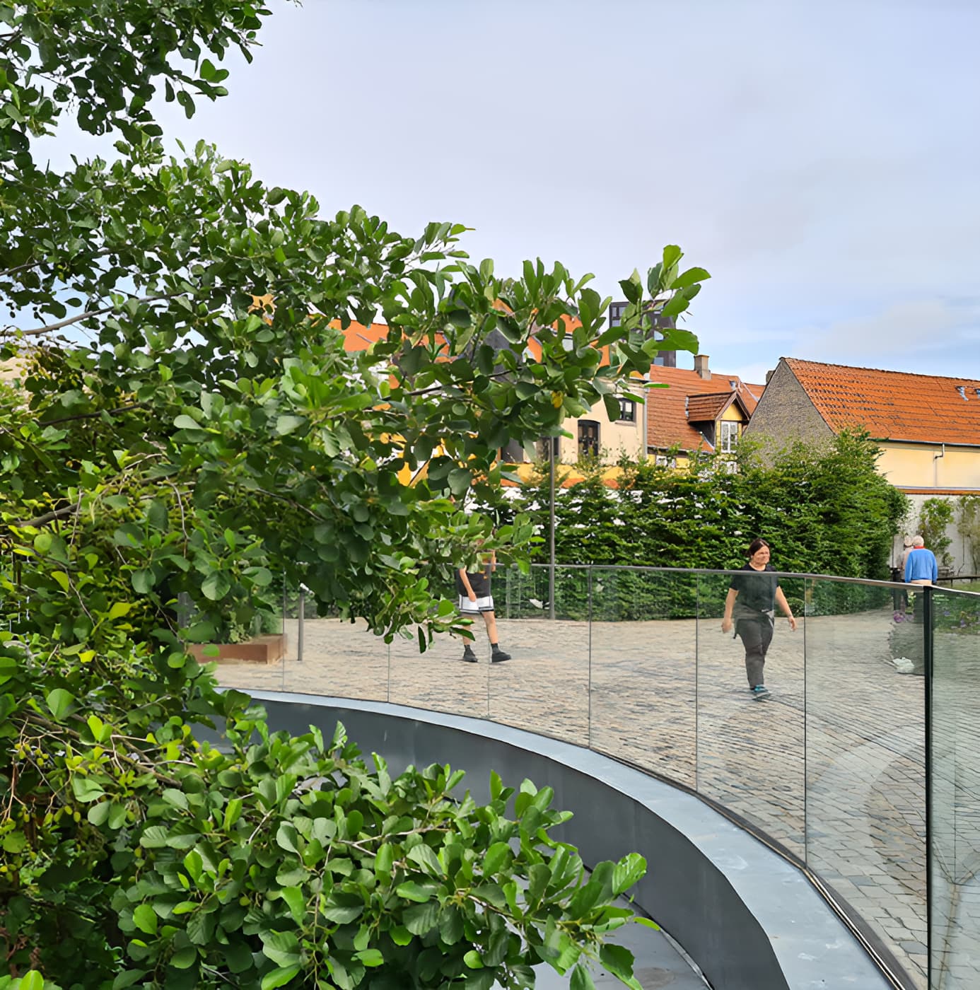 Curved glass railing surrounding a cobblestone plaza with lush green trees and traditional European buildings in the background