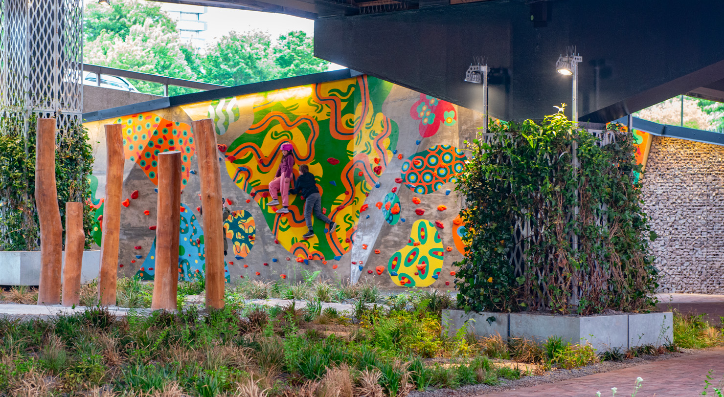 Two people climb a large outdoor bouldering wall covered in a vivid abstract mural, set within a landscaped urban space featuring timber posts, planted borders, and climbing ivy.
