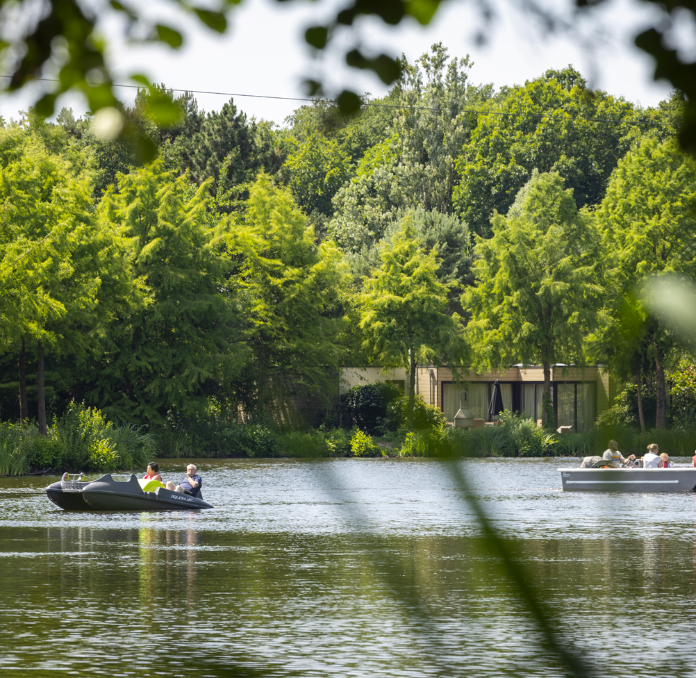 Two small leisure boats cross a calm lake surrounded by dense summer woodland, with a contemporary timber pavilion visible on the far bank, suggesting a well-managed outdoor leisure environment.