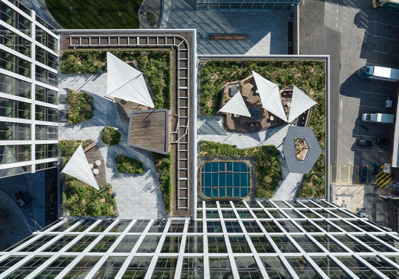 Aerial view of a lush rooftop garden with white sail shades, wooden decking, green planting and a small pool atop a city building