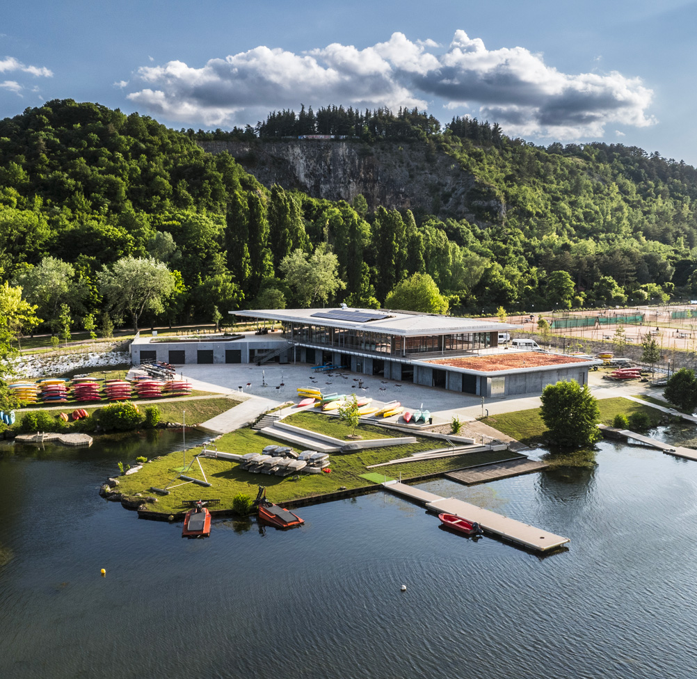 Aerial view of a lakeside outdoor sports and leisure centre with kayaks and canoes arranged on the quayside, solar panels on the rooftop, and a wooded hillside with a rocky escarpment rising beyond.