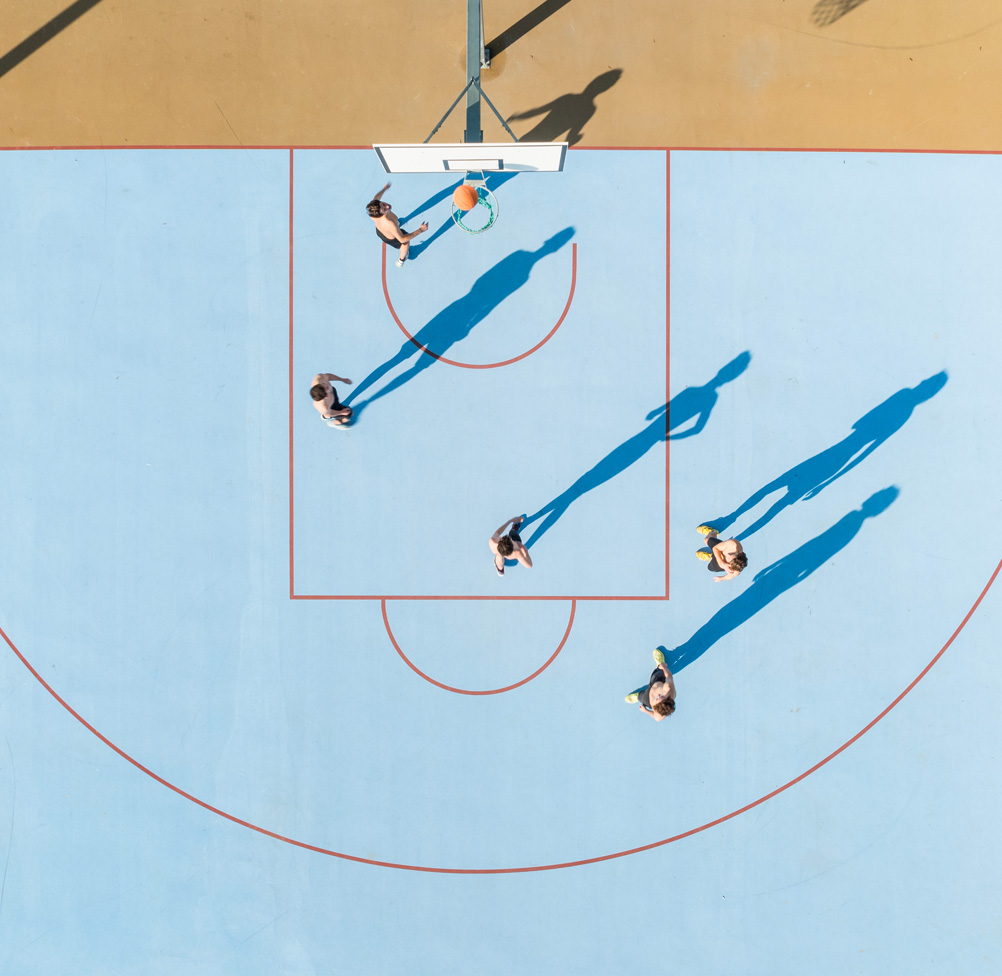 Overhead drone view of five players in a basketball game on a vivid blue and sand-coloured outdoor court, with long shadows cast across the painted surface.