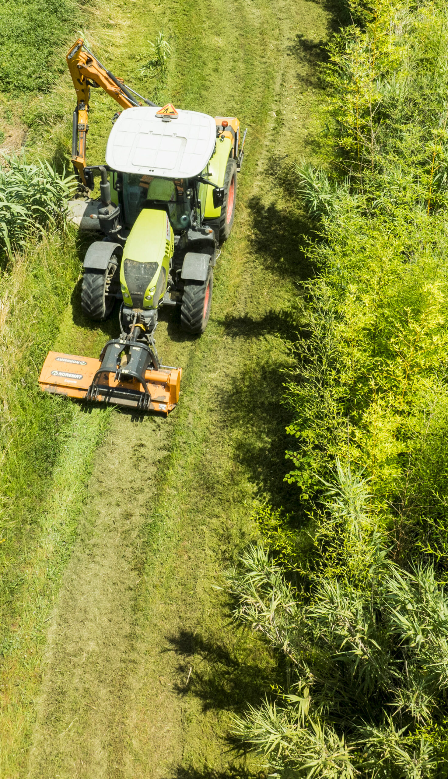Aerial view of a Claas tractor fitted with a front flail mower and rear hedgecutter arm working along a managed green corridor between dense scrubland and reed vegetation.