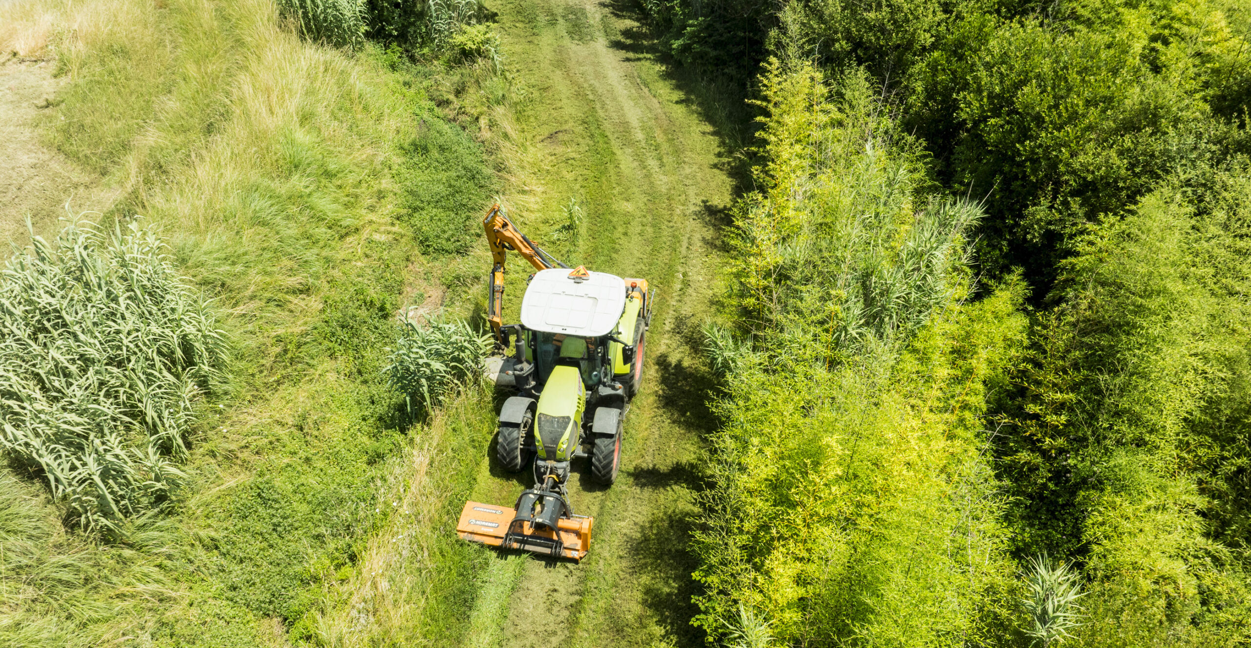Aerial view of a Claas tractor fitted with a front flail mower and rear hedgecutter arm working along a managed green corridor between dense scrubland and reed vegetation.