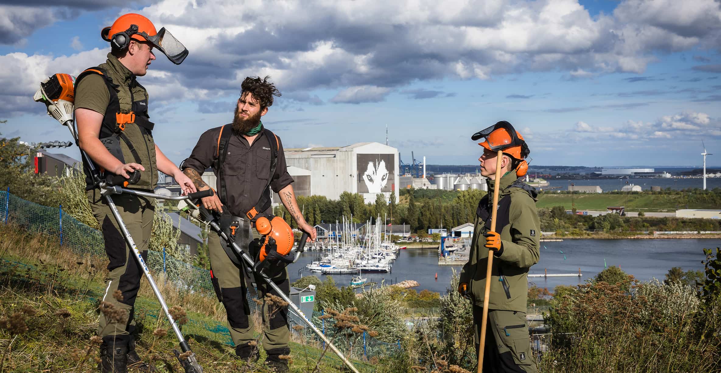 Three idverde grounds maintenance workers with orange helmets and STIHL equipment on a hillside overlooking a marina and harbour.