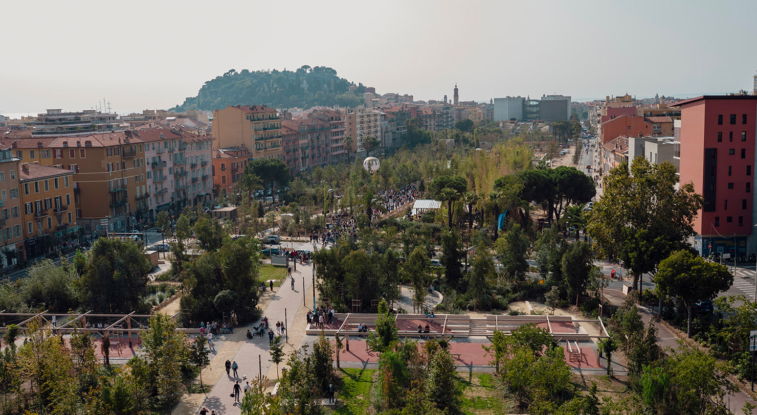 Aerial view of a newly landscaped urban park in Nice, with tree-lined walkways, seating areas and play spaces woven through a dense city fabric, drawing large crowds on opening day with the Colline du Château visible in the background.