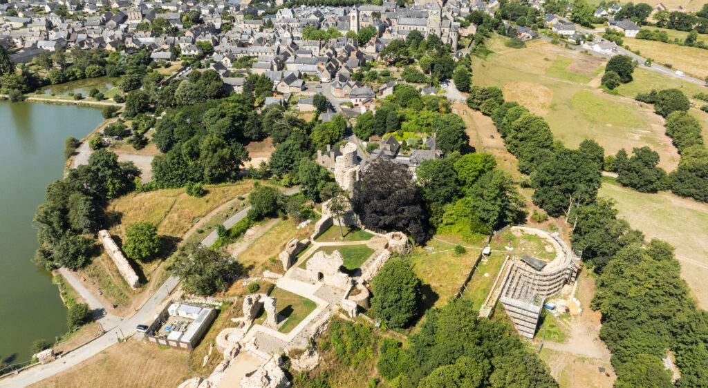 Aerial view of a medieval castle ruin set within managed green grounds alongside a French market town, with restoration scaffolding visible on one tower and a lake bordering the site, illustrating the maintenance of heritage landscapes.