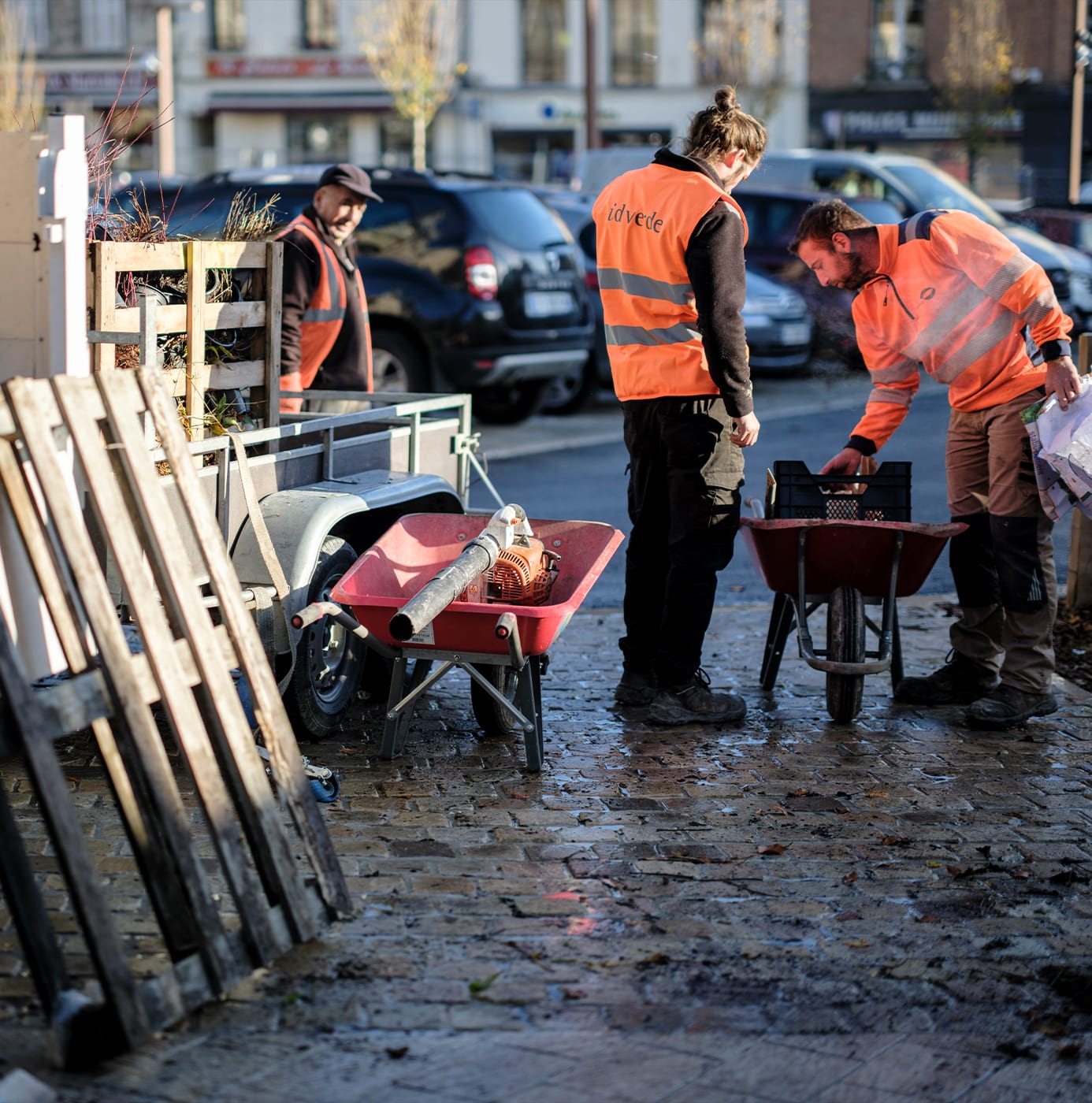 idverde workers in orange hi-vis vests managing landscaping equipment and wheelbarrows on a wet cobblestone street.
