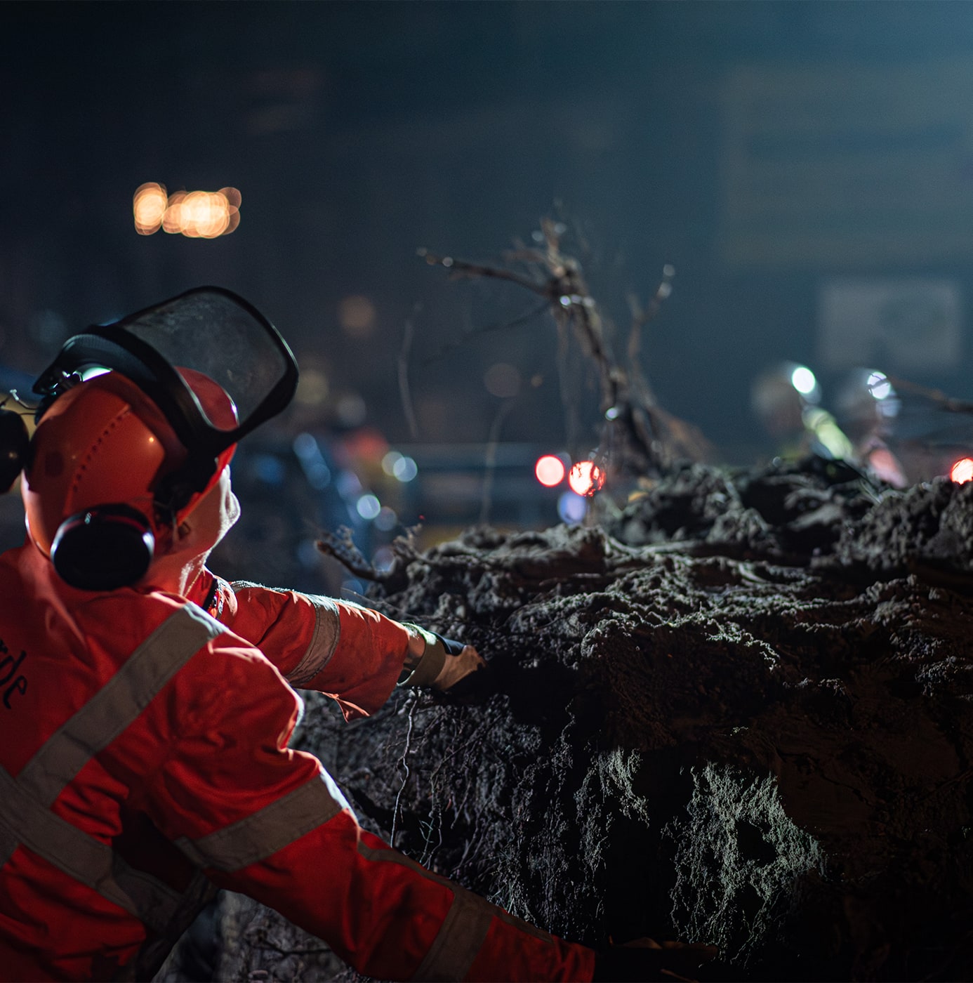 idverde worker in red PPE and face shield operating a chainsaw on a fallen tree at night