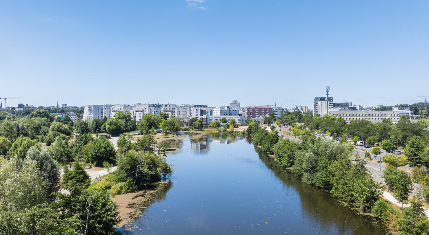 A tree-lined urban river corridor runs through a French city in summer, with managed green banks, public pathways and residential buildings reflected in the calm water under a clear blue sky.