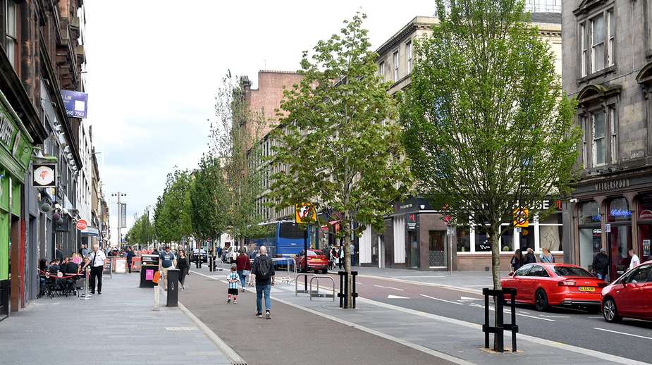 Tree-lined urban street with young deciduous trees, wide pedestrian pavements, and mixed-use retail buildings in a UK city centre.